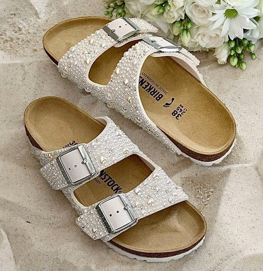 White Birkenstock Arizona bridal sandals with pearls, delicate pearl hearts and crystal accents, displayed on sand beside white flowers.