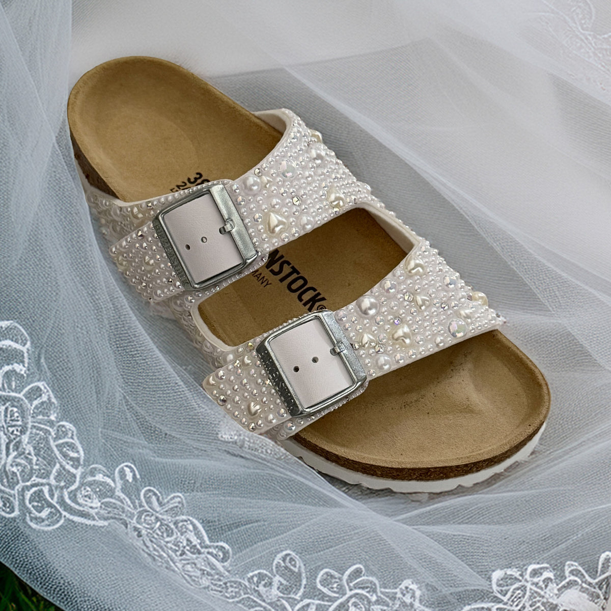 White Birkenstock Arizona sandal with pearls, delicate pearl hearts and crystal accents, displayed on embroidered bridal tulle.