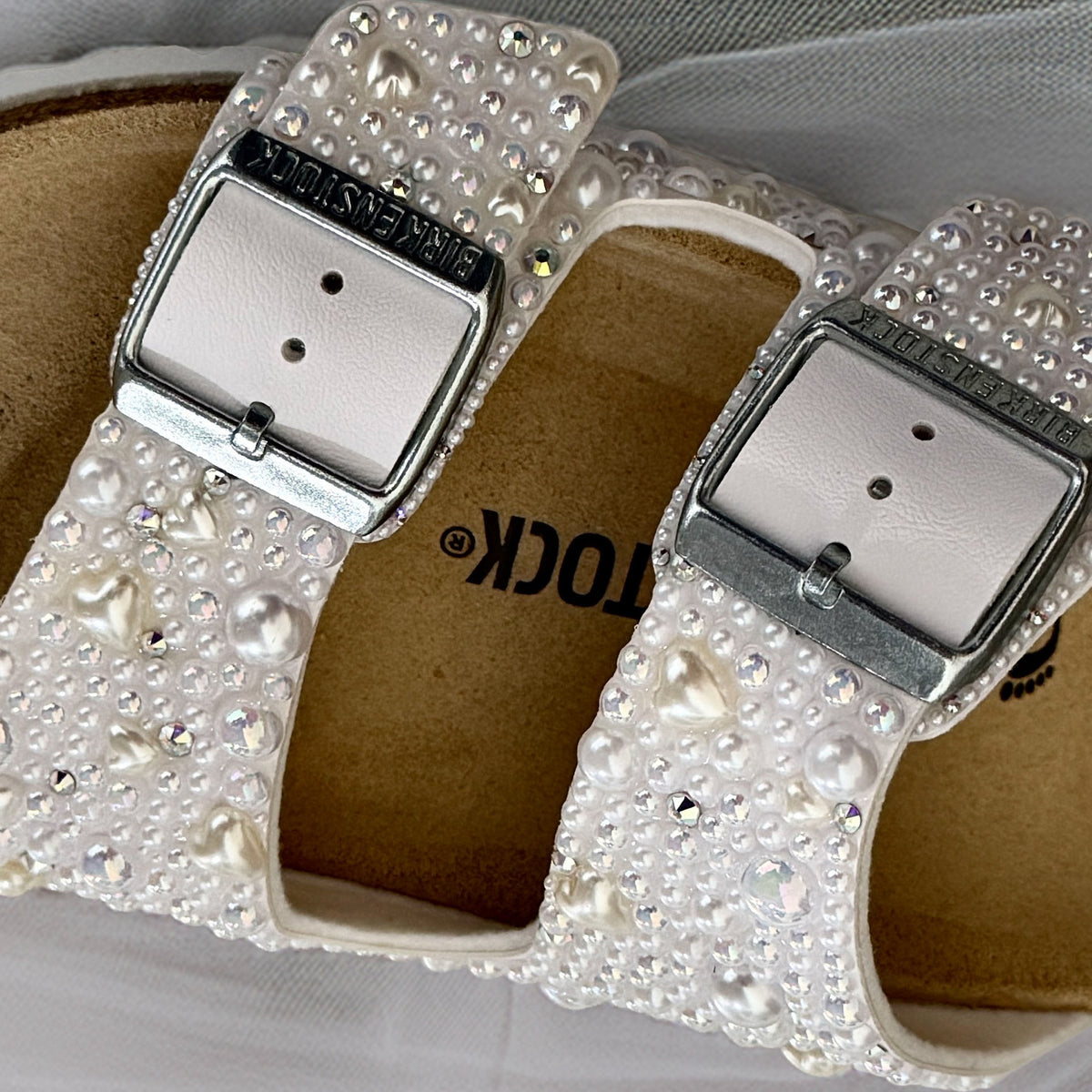 Close-up of silver buckles and white straps decorated with pearls, delicate pearl hearts and crystal accents on a Birkenstock Arizona sandal.